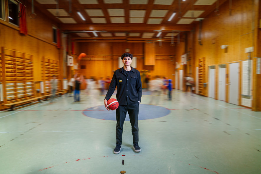 Monjed med basketboll i gymnastiksal. Barn rör sig i bakgrunden.